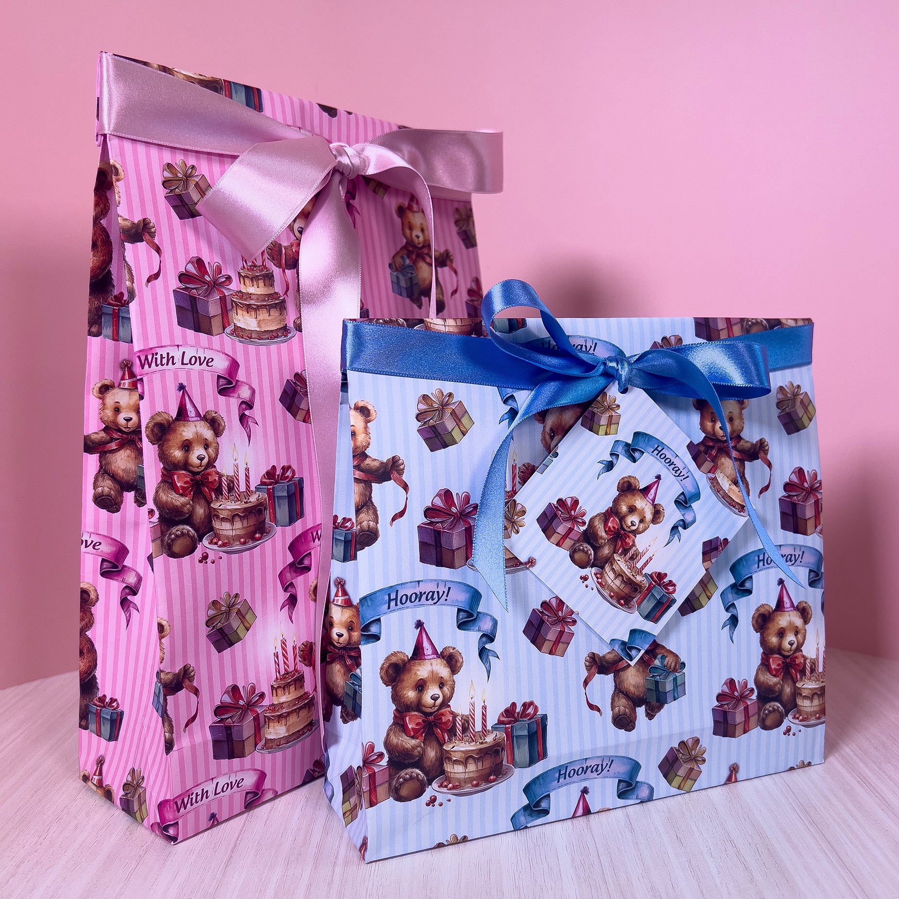 Two decorative gift bags with teddy bear and birthday cake designs on a pink background.